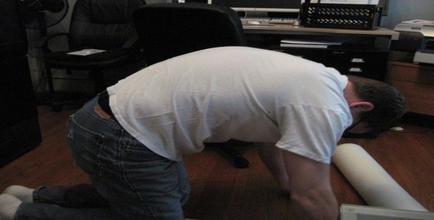 A man kneeling on the floor in an office. He is wearing a white t-shirt and blue jeans. His head is down and his hands are resting on his knees. In front of him there is a roll of white paper. The office has a desk with a computer monitor keyboard and other office supplies. There is a black office chair in the background. The floor is made of wood and the walls are painted white.
