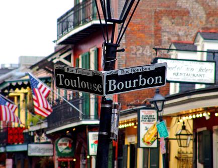 A street sign on a pole in the middle of a busy street. The sign is black and white and reads "Toulouse Bourbon" in French. The street is lined with buildings on both sides and there are American flags hanging from the balconies. The buildings appear to be old and dilapidated with boarded up windows and boarded up doors. There are also several signs and advertisements on the buildings. The sky is overcast and the overall mood of the image is gloomy.