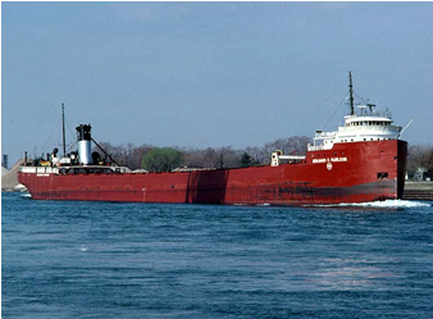 A large red cargo ship sailing on the water. The ship has a white superstructure with a red stripe running along the side and a large antenna on top. There are several smaller antennas and other equipment on the deck of the ship. The sky is blue and the water is calm with small waves visible on the surface. In the background there are trees and buildings visible.