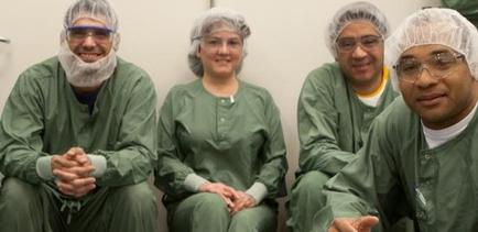 Four people two men and two women sitting in a row and smiling at the camera. They are all wearing green surgical gowns and white hairnets on their heads. The man on the left is wearing a blue surgical mask and goggles the man in the middle is wearing an orange shirt and the woman on the right is wearing glasses. They appear to be in an operating room with a white wall in the background. All four people are sitting with their hands clasped together in front of them.