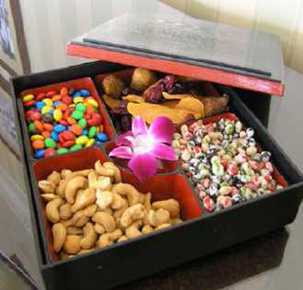 A black rectangular box with a red lid. Inside the box there are various types of nuts and candies. On the left side there is a small red container filled with colorful candies and on the right side it is filled with a variety of nuts including cashews almonds and peanuts. In the center of the box is a pink flower adding a pop of color to the scene. The box appears to be sitting on a wooden table with a white wall in the background.
