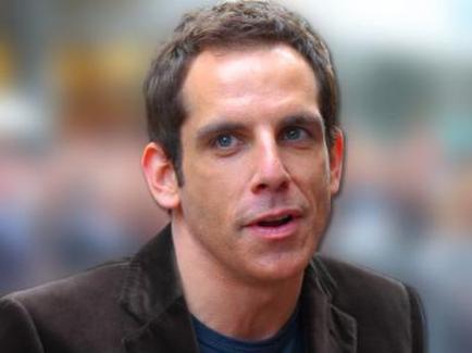 A close-up of a man's face. He appears to be in his late 40s or early 50s with short dark hair and a slight stubble. He has a serious expression on his face and is looking off to the side with a slight smile. He is wearing a dark brown jacket over a blue t-shirt. The background is blurred but it seems to be an outdoor setting with people in the distance.