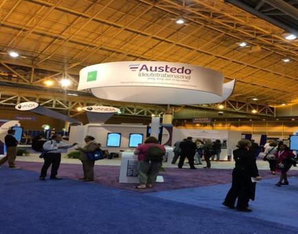 A large exhibition hall with a blue carpeted floor and a high ceiling with wooden beams. In the center of the hall there is a large white banner with the word "busted" written in bold black letters. Above the banner there are several smaller banners with the same word written in smaller black letters indicating that this is a trade show booth. There are several people gathered around the booth some of them are interacting with each other while others are looking at the banners. The booth appears to be set up for an event or trade show.