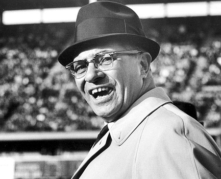A black and white photograph of a man wearing a hat and glasses. He is standing in front of a large crowd of people in a stadium. The man appears to be in his late 60s or early 70s with a wide smile on his face. He has short dark hair and is wearing a collared shirt and a tie. The background is blurred but it seems to be a large arena with rows of seats. The image is likely taken at a sporting event or a rally.