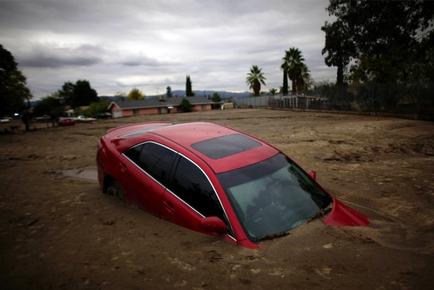 A red car that has been flipped over in a muddy area. The car appears to be a sedan or a hatchback with the hood crumpled and the windshield shattered. The ground is covered in mud and debris and there are trees and houses in the background. The sky is cloudy and the overall mood of the image is one of destruction and chaos.