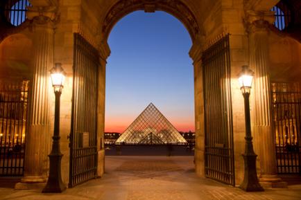 An archway with two lanterns on either side. The archway is made of stone and has intricate carvings on the top and sides. The lanterns are turned on casting a warm glow on the archway. In the background there is a beautiful view of the Louvre Pyramid in Paris France. The sky is a deep blue and the sun is setting creating a warm orange glow. The pyramid is the focal point of the image with its distinctive triangular shape and intricate details.