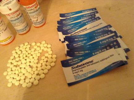 A pile of white pills on a beige countertype. On the left side of the image there are several prescription bottles of different sizes and colors including orange white and blue. Next to the bottles there is a pack of suboxide tablets. The tablets are arranged in a scattered manner with some of them overlapping each other. The pack of tablets has a blue label with white text and numbers on it. The background is blurred but it appears to be a kitchen countertype with other items scattered around.
