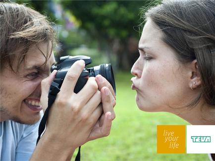 A man and a woman taking a photo together with a DSLR camera. The man is holding the camera in his hands and the woman is kissing him on the cheek. They are standing in a park or garden with trees and grass in the background. The woman has her eyes closed and the man is looking at her with a smile on his face. The image appears to be taken during the day.