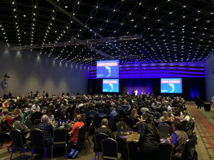 A large conference room with a high ceiling and rows of chairs. The room is filled with people sitting at tables facing a stage where a man is giving a presentation. The stage is decorated with blue and purple lights and there are two large screens on either side of the stage. The audience is attentively listening to the presentation. There are cameras on the stage suggesting that the event is taking place in a professional setting. The overall atmosphere of the room is formal and professional.