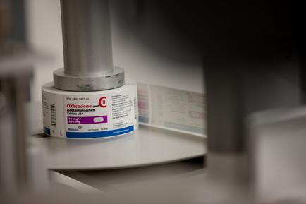 A white cylindrical container with a blue label on it. The label has the brand name "Oxycodone" written in bold black letters at the top. Below the label there is a description of the product which is written in smaller black letters. The container is placed on a white surface and there is another label visible in the background. The background is blurred but it appears to be a laboratory or laboratory setting.