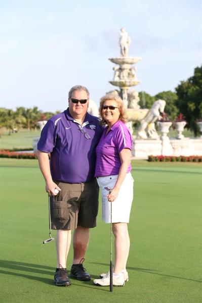 A man and a woman standing on a golf course. They are both holding golf clubs and are smiling at the camera. The man is wearing a purple polo shirt khaki shorts and black shoes. He has a pair of sunglasses on and is holding a golf club in his left hand. The woman is also wearing a pink shirt and white shorts. In the background there is a fountain with statues and flowers. The sky is blue and there are trees in the distance.
