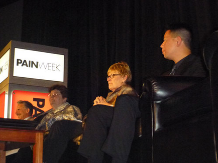 A group of three people sitting on a stage with a black curtain in the background. On the left side of the stage there is a podium with a sign that reads "PAIN WEEK". On the right side there are two men sitting in black leather chairs facing each other. The man on the left is wearing a suit and tie and appears to be speaking into a microphone. The woman in the middle is sitting with her hands clasped together and is looking towards the audience. The other two men are listening attentively. All three people are wearing casual clothing and appear to be engaged in a discussion.