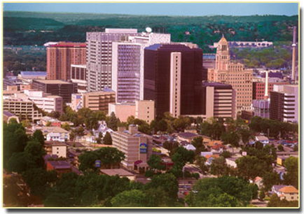 An aerial view of a city with tall buildings and skyscrapers. The city appears to be a mixed-use development with a variety of modern and high-rise buildings. The buildings are of different sizes and shapes with some taller and others smaller. In the center of the image there is a tall modern building with a dome on top. To the right of the building there are several smaller buildings with a clock tower. The sky is blue and there are trees and greenery scattered throughout the city. The image is taken from a high vantage point looking down on the city below.