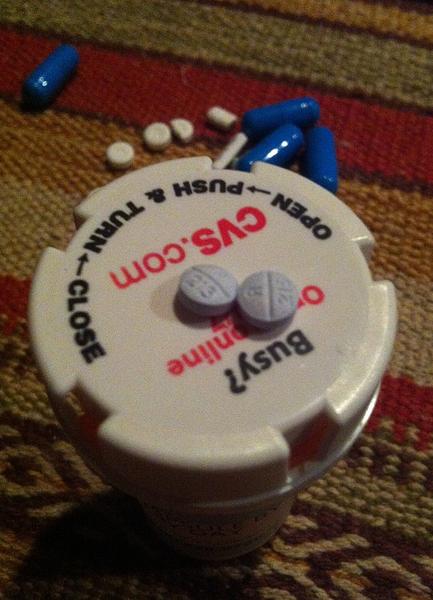 A white pill bottle with a red label on it. The label has the words "CVS.com" written in red and black letters. Below the label there are two white pills one of which is open and the other is closed. There are also two blue pills scattered around the bottle. The bottle appears to be lying on a striped carpeted floor.