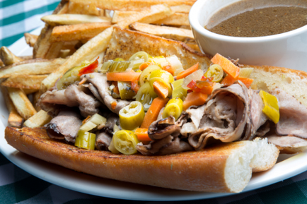A plate with a sandwich and french fries. The sandwich is made with a long toasted bun and is filled with sliced meat vegetables and pickles. The meat appears to be roast beef and the vegetables include carrots celery and onions. The fries are golden and crispy. On the right side of the plate there is a small bowl of brown sauce. The plate is white and the background is a green and white checkered tablecloth.