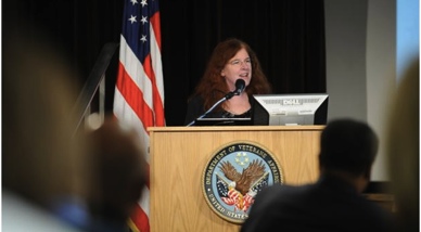 A woman standing behind a wooden podium with the seal of the United States of America Department of Veterans Affairs on it. She is speaking into a microphone and appears to be giving a speech or presentation. The background is dark and there is an American flag on the left side of the image. The woman has long red hair and is wearing a black blazer. There is an audience sitting in front of the podium.