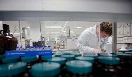 A young man in a laboratory setting. He is wearing a white lab coat and glasses and is working on a piece of equipment. He appears to be focused on his work. In front of him there are several rows of blue-colored jars with lids. On the left side of the image there is a large brown bottle with a label that reads "High Technology". In the background we can see other laboratory equipment and equipment. The overall atmosphere of the laboratory is clean and organized.