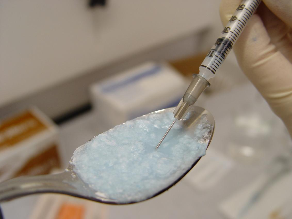 A spoon filled with a light blue substance likely an opioid or another drug and a hand holding a syringe. The syringe drawing up the substance.  The hand is wearing a white safety glove. The background is blurred but it appears to be  laboratory setting.