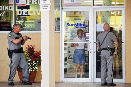 Two police officers standing in front of a store front. The store has a sign that reads "Sunshine Pharmacy" and there is a picture of a woman in a blue dress on the glass door. The officers are wearing grey uniforms and carrying guns. One officer is holding a gun and appears to be directing the other officer. There are potted plants and flowers in the background.