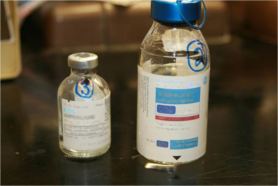 Two small vials labeled 3 (containing Mepivacaine on the left) and 2 (containing Visipaque on the right). The vials are clear with screw-on tops. Their labels are both blurry and faded suggesting that the are old and have been heavily used. A dark scuffed up table is visible in the background.