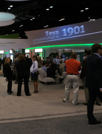 A large exhibition hall with a group of people gathered around a booth. The booth has a sign that reads "tea 1901 established in 1901" and is lit up with green lights. There are several people walking around the booth some of them are engaged in conversation while others are sitting on sofas. The floor is covered in a gray carpet and there are several other booths visible in the background. The overall atmosphere of the exhibition hall appears to be busy and bustling.