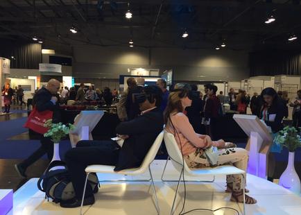 A large exhibition hall with a high ceiling and multiple levels. There are several people in the hall some standing and some walking. In the center of the image there are two people sitting on white chairs facing each other. The person on the left is wearing a black suit and appears to be engaged in a conversation with the person in the middle who is also wearing a virtual reality headset. On the right there is a woman wearing a pink top and beige pants who seems to be listening intently to the conversation. In front of the two people on the chairs is a white table with a vase of green plants on it. The background shows other booths and tables with various items on display.