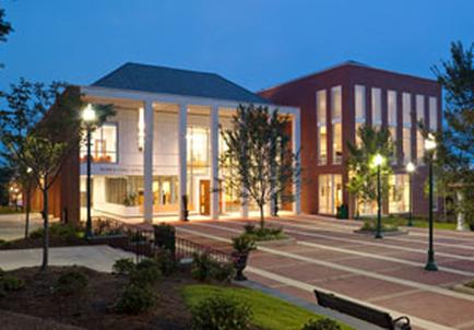 A large modern building with a red brick exterior and white columns. The building is located in a park-like setting with a paved walkway in front of it. The walkway is lined with trees and shrubs and there are street lamps on either side. The sky is dark indicating that it is either dusk or early morning. In front of the building there is a bench suggesting that the photo was taken at night. The overall atmosphere of the image is peaceful and inviting.