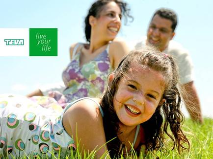 A young girl lying on the grass with her family in the background. The girl is smiling and looking at the camera with a big smile on her face. She is wearing a colorful dress with a floral pattern and has long curly hair. Behind her there is a man and a woman both of whom are also smiling and appear to be happy. The background is a clear blue sky and the grass is green. On the top left corner of the image the words "Live your life" are written in white text.