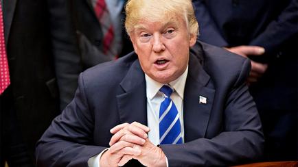 Former US President Donald Trump sitting at a desk with his hands clasped together in front of him. He is wearing a dark suit and a blue tie. He appears to be deep in thought with a serious expression on his face. In the background there are other people wearing suits and ties suggesting that he is in a formal setting. The image appears to have been taken in a government or government building.