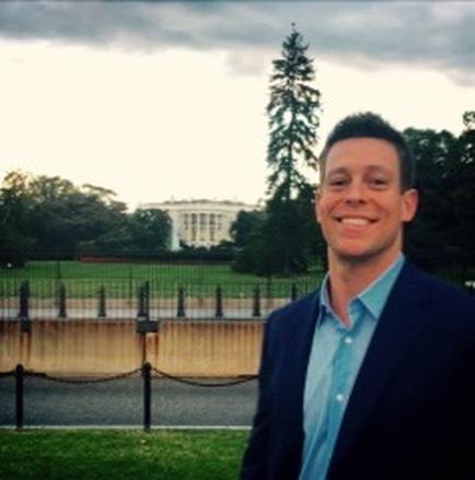A portrait of a young man standing in front of the White House. He is wearing a blue blazer and a light blue shirt. He has short dark hair and is smiling at the camera. The background of the image shows the white house which is visible in the distance. The sky is cloudy and there are trees and a fence in the foreground.
