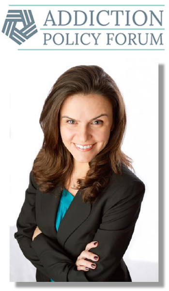 A professional headset of a woman. She is wearing a black blazer and a turquoise blouse. She has long brown hair and is smiling at the camera. Her arms are crossed in front of her and she appears to be confident and professional. The background is white and there is a logo for the Addiction Policy Forum on the top left corner.