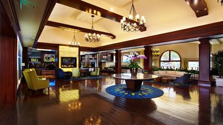 A luxurious lobby of a hotel. The lobby has a high ceiling with wooden beams and a large chandelier hanging from it. The floor is made of hardwood and there is a round wooden coffee table in the center of the room. Around the coffee table there are several colorful armchairs and sofas in different colors and styles. On the left side of the image there is an entertainment center with a flat-screen TV mounted on the wall. The walls are painted in a warm yellow color and there are large windows that let in natural light. The overall atmosphere of the lobby is inviting and inviting.