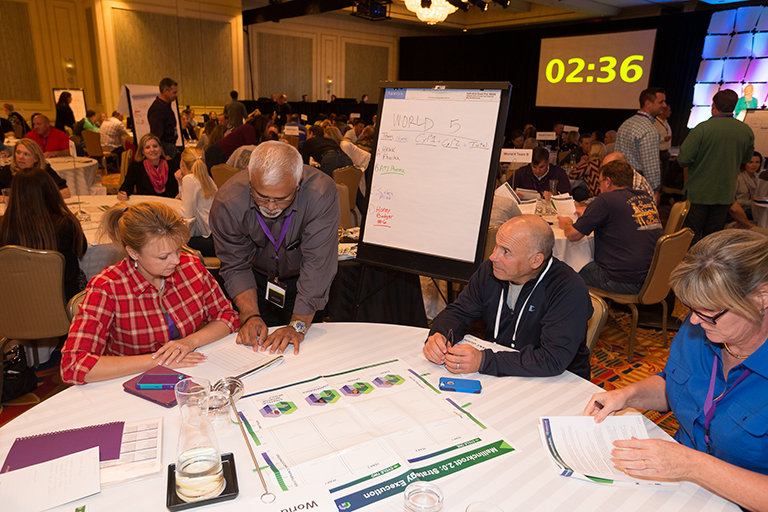 A group of people sitting at a table in a conference room. There are four people in the image two men and two women all of whom appear to be engaged in a discussion. The table is covered with a white tablecloth and there is a large whiteboard behind them. A screen in the background has "02:36" written on it. The people are looking at the whiteboard and seem to be working on a project or presentation. There is a chandelier hanging from the ceiling and a large screen in the background. The room is filled with other people some of whom are also sitting at tables and some are standing.