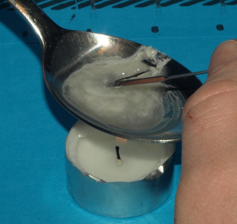 A person's hand holding a silver spoon over a small white tea candle. The spoon is filled with a white substance likely an opioid or another drug. The candle is lit and the background is a blue surface. On the right side of the image a person's hand appears to be stirring the substance with what appears to be a metal needle or nail.