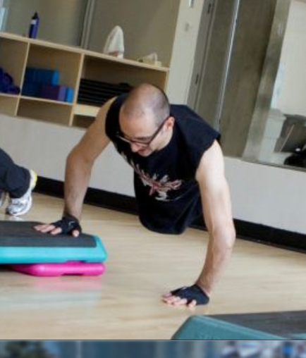 A man in a gym performing a push-up exercise. He is wearing a black tank top with a graphic design on it and black gloves. His arms are stretched out in front of him and his legs are bent at the knees. He appears to be in the middle of a plank position with his hands on the ground and his feet flat on the floor. In the background there are shelves with exercise equipment and a mirror on the wall.