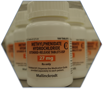 A white medicine bottle with a white cap. The orange label on the bottle reads "Methylphenidate Hydrochloride Extended-Release Tablets USP 27 mg Rx only". The label also reads: "Pharmacist: Dispense the Medication Guide provided separately to each patient." In the background there are other white medicine bottles with orange labels in a triangle formation. The image appears to be in a hexagon.