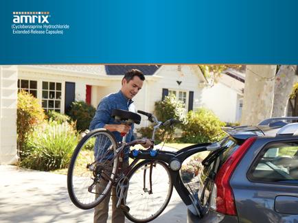 A man standing next to a car with a bicycle attached to the back of it. The man is wearing a blue shirt and khaki pants and is holding the handlebars of the bicycle with both hands. He is smiling and appears to be loading the bicycle onto the car. The car is parked on a driveway in front of a house with a white exterior and a red door. There are plants and trees in the background. The image is accompanied by the logo of Amrix a company that provides extended-release capsules.