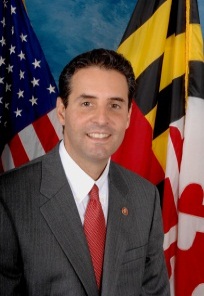 A portrait of a man standing in front of two flags - the United States and the Maryland state flag. The man is wearing a gray suit and a red tie and has a small button on his lapel. He is smiling and looking directly at the camera.