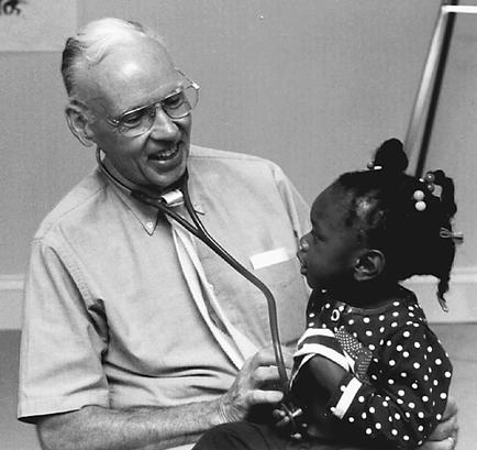 A black and white photograph of an elderly man with a stethoscope around his neck sitting on a chair and talking to a young girl. The man is wearing a collared shirt and glasses and appears to be in his late 60s or early 70s. The girl is sitting on the man's lap and is looking up at him attentively. She has her hair tied up in a bun and is wearing polka-dot dress. The background is a plain wall with a sign that reads "No. 6". The image appears to have been taken in a hospital or clinic setting.