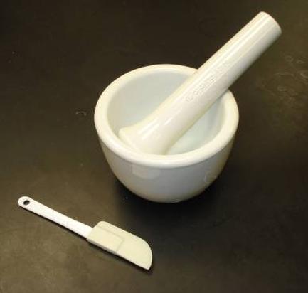 A white mortar and pestle on a black surface. The mortar is made of ceramic and has a round base with a handle on one side. The pestle is also white and appears to be made of porcelain. On the other side of the mortar there is a small white spatula with a curved handle. The spatula is resting on the surface next to the mortar.