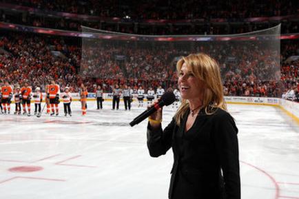 A woman holding a microphone and standing on the ice of an ice hockey rink. She is wearing a black blazer and appears to be speaking to the crowd. In the background there is a group of hockey players from the Philadelphia Flyers team standing in formation on the rink. The players are wearing orange and white jerseys with the team's logo on the front. The rink is surrounded by a large crowd of spectators in the stands. The image appears to have been taken at a hockey game.