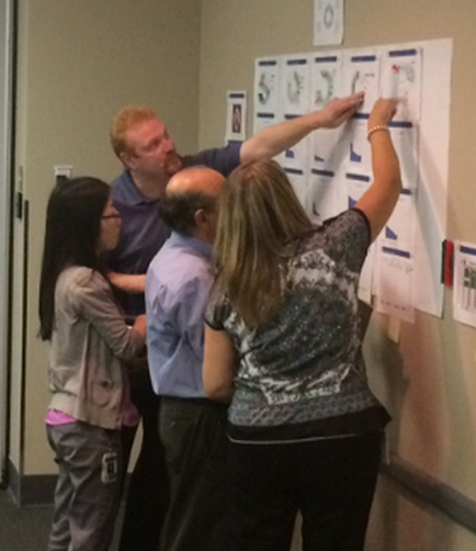 A group of five people standing in a room with a whiteboard on the wall. The whiteboard is covered with multiple posters and charts. There are four people in the image three men and two women all of whom appear to be engaged in a discussion. The man on the left is pointing to one of the posters on the whiteboard while the other two are looking at it intently. The woman on the right is wearing a gray blouse and black pants and the man in the middle is holding a pen and appears to be writing on a piece of paper. The other three people are standing around the board looking at the posters. There is a door visible in the background.