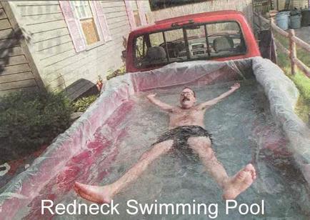 A man lying in a redneck swimming pool. He is wearing black shorts and appears to be in the middle of a swim. His arms are stretched out to the sides and his legs are bent at the knees. The pool is covered in water and there is a red truck parked next to it. The background shows a house with a wooden fence and some trash cans. The text on the image reads "Redneck Swimming Pool."