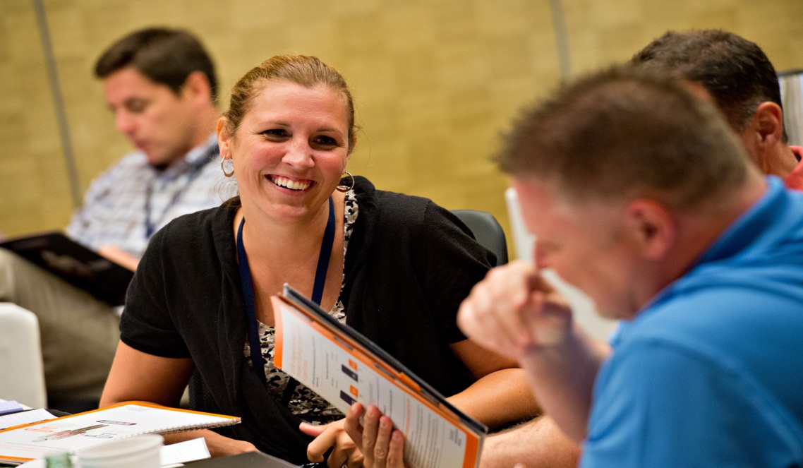 A group of people sitting at a table in a conference room. There are four people in the image three men and one woman. The woman is in the center of the image and is smiling. She is wearing a black top and has a lanyard around her neck. The man on the left is holding a folder. Two people are sitting on either side of the table and are looking at the folder. There are some papers on the table in front of them. The background is blurred but it seems like the room is well-lit with natural light coming in from the windows.