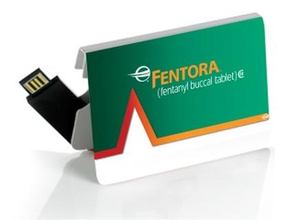 A USB flash drive with a green card attached to it. The card has the word "mentor" written in white capital letters on the top left corner followed by the company's logo. Below the logo there is a red arrow pointing upwards indicating that the flash drive can be used to transfer data from one device to another. The flash drive is attached to a white stand with a black connector on the left side. The background is white.