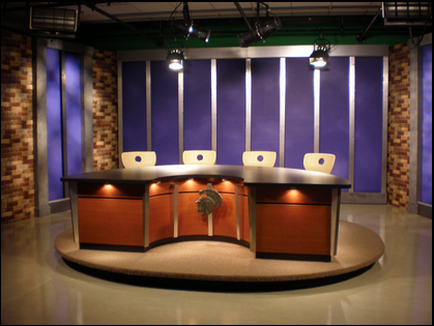 A modern TV studio set with a large wooden desk in the center. The desk has a curved top and four white chairs around it. The walls are made of brick and there is a large window on the right side of the desk with blue curtains. Above the desk there are three spotlights hanging from the ceiling. The floor is made of concrete and the overall color scheme of the room is warm and inviting. The overall design of the studio is sleek and contemporary.