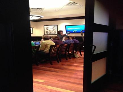 A group of people sitting at a long table in a room with wooden flooring. The room appears to be a conference room or a meeting room as there is a large screen on the wall in the background. The people are sitting in rows of wooden chairs facing the screen and appear to be engaged in a discussion. There is a woman standing at the front of the room holding a microphone and speaking to the group. On the left side of the image there are several framed pictures hanging on the walls. The image is taken from a doorway looking out onto the room.