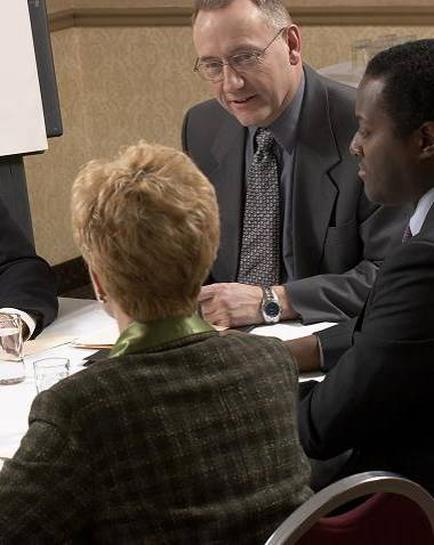 A group of three people sitting around a table in a meeting room. The person in the center is a man wearing a suit and tie and he appears to be speaking to the two people on either side of him. The woman on the left is a middle-aged woman with short blonde hair wearing a green blazer and a green scarf. The man on the right is a young African-American man also wearing a black suit. They are all looking at each other attentively and appear to be engaged in a conversation. There is a projector screen in the background suggesting that the meeting is taking place in a professional setting.
