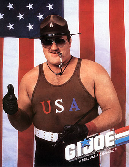 A promotional poster for the movie "G.I. Joe: A Real American Hero." It features a man standing in front of an American flag. The man is wearing a brown tank top with the word "USA" written on it in red white and blue letters. He is also wearing a black cowboy hat sunglasses and a lanyard around his neck. He has a mustache and is giving a thumbs up with his right hand. The background is white with red and blue stripes. The title of the movie is written in blue letters at the bottom of the image.