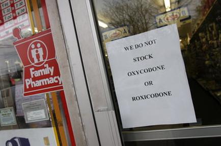 A sign on the door of a Family Pharmacy store. The sign is white with black text that reads "We do not stock oxycodone or roxicodone". The store's name is written in bold letters at the top of the sign. Below the sign there is a red sign with the words "Family Pharmacy" written in white letters. The background of the image shows the store's windows and shelves with various items for sale.